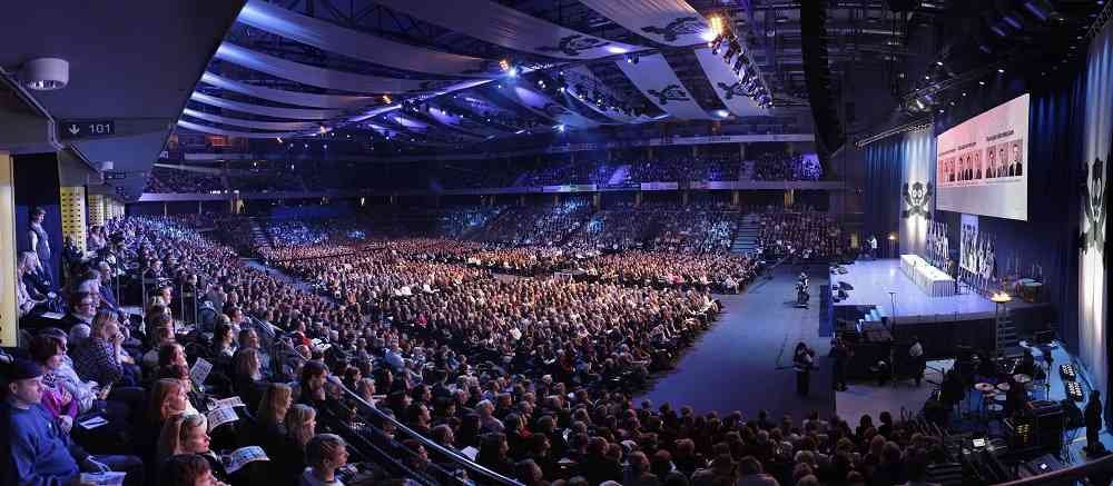 A packed arena audience facing a lit stage with large screens.